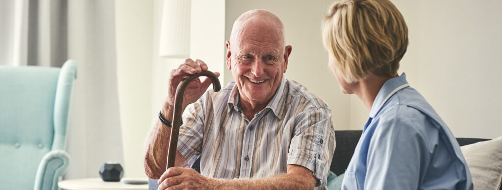 Ein älterer Mann mit Gehstock sitzt entspannt neben einer Pflegefachperson und lächelt – eine Szene, die die Bedeutung von Stressbewältigung für pflegende Angehörige unterstreicht. Das Bild symbolisiert die wertvolle Unterstützung im Alltag und verweist auf das Engagement der IAHA (International Association for Healthy Aging) für mentale Gesundheit und Entlastung in der häuslichen Pflege.