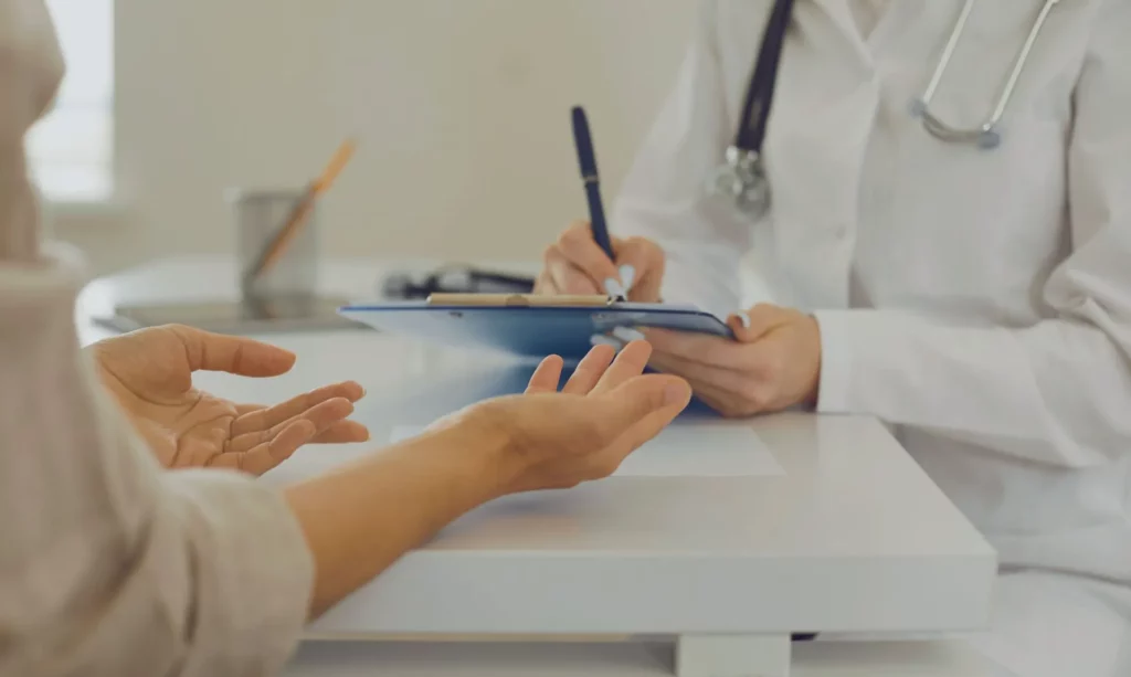 Ein Arzt schreibt Notizen auf ein Klemmbrett, während eine Person ihm gegenübersitzt und gestikulierend Fragen stellt. Die Szene verdeutlicht die Abrechnung im Gesundheitswesen. Die IAHA (International Association for Healthy Aging) unterstützt Patient:innen und pflegende Angehörige bei der Abrechnung von Pflegeleistungen mit individueller Beratung und praxisnahen Hilfestellungen.