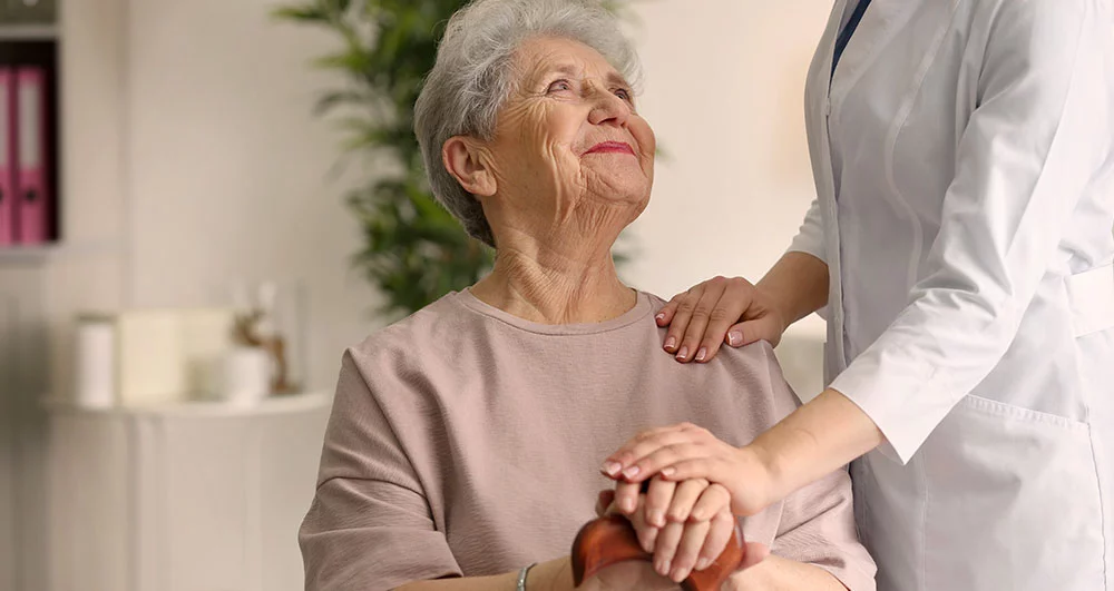 Eine ältere Frau sitzt lächelnd auf einem Stuhl und schaut dankbar zu einer Pflegekraft, die ihr beruhigend die Hände hält. Die Szene symbolisiert Fürsorge und Unterstützung für pflegende Angehörige. Die IAHA (International Association for Healthy Aging) bietet umfassende Beratung und praxisnahe Hilfe, um die Lebensqualität pflegende Angehörige zu sichern und ihre Betreuung zu verbessern.